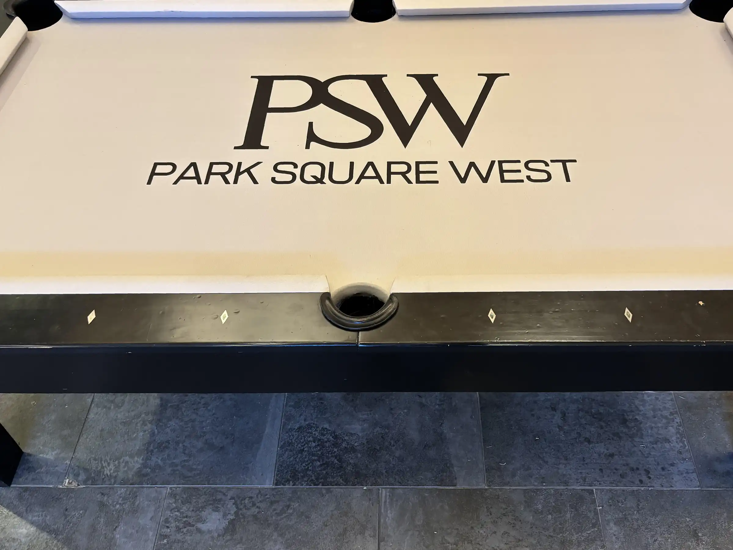 A pool table with a "PSW" logo and "Park Square West" text. The table has a black frame and a white felt surface.