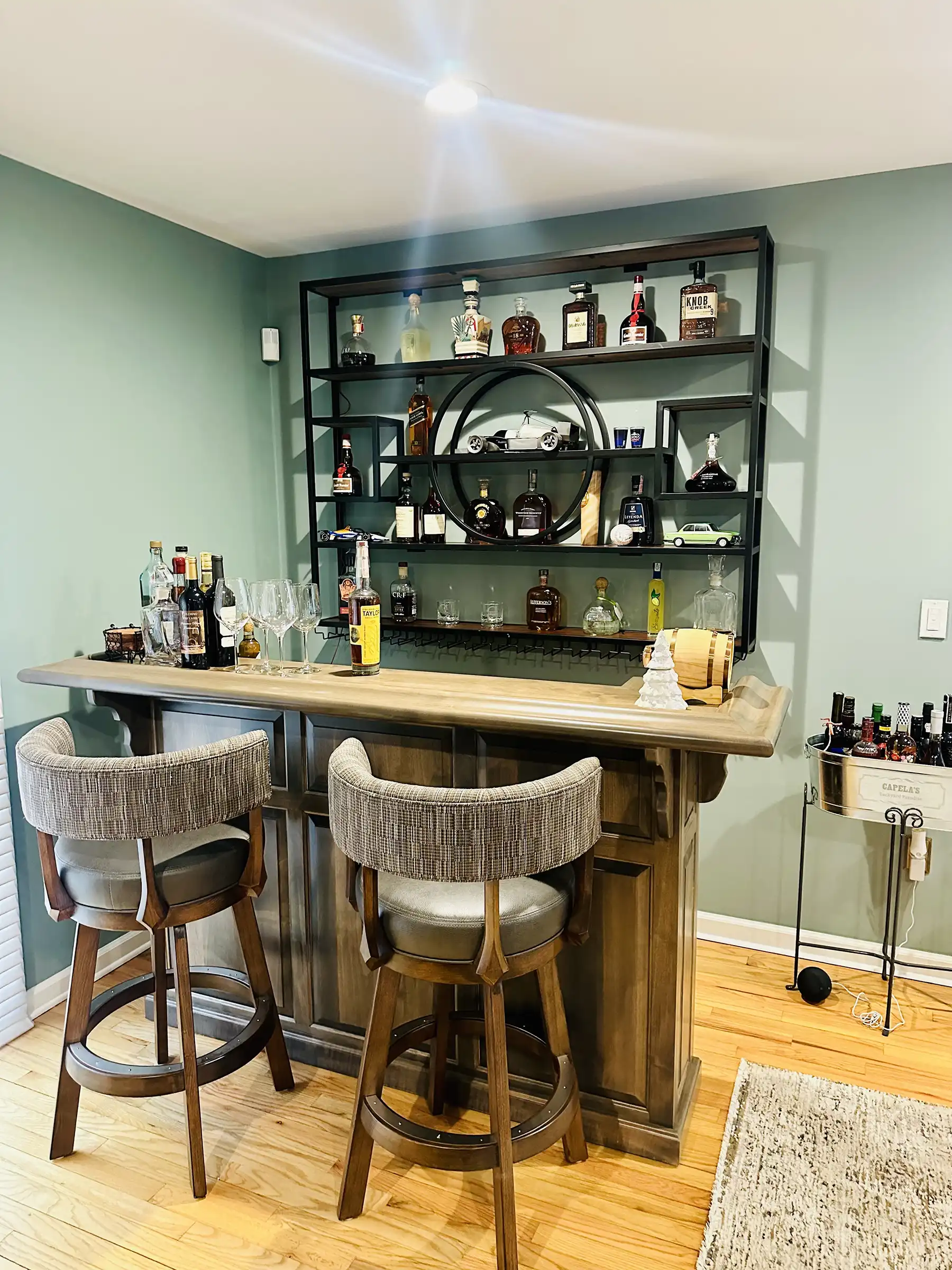 Modern home bar with a unique metal shelf showcasing various spirits. The bar features a wooden countertop, two comfortable bar stools, and a small bar cart for additional storage.