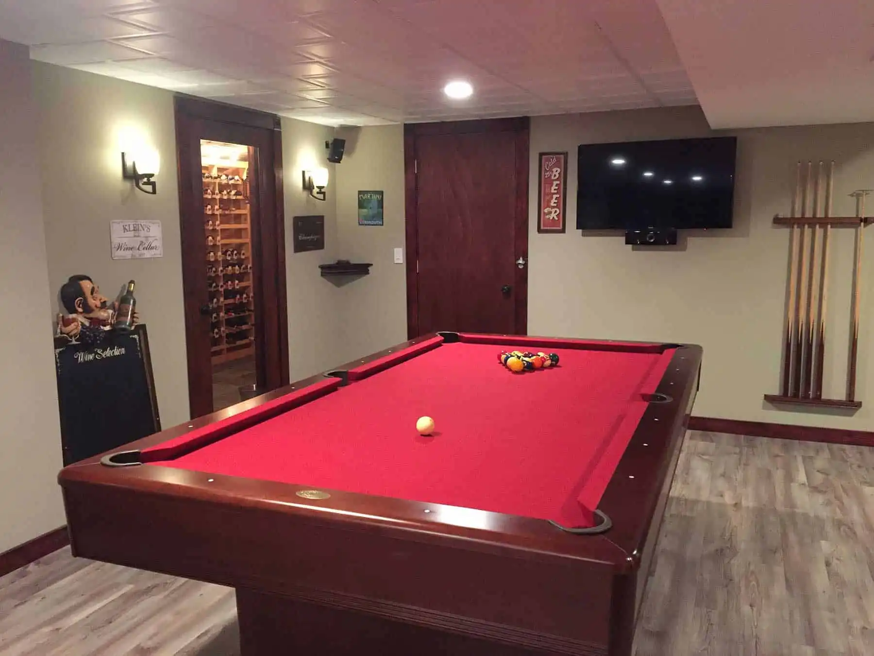 Basement game room featuring a mahogany pool table with red felt, a cue rack, built-in wine cellar, wall-mounted TV, and cue rack, decorated with vintage beer signs and wood-look flooring