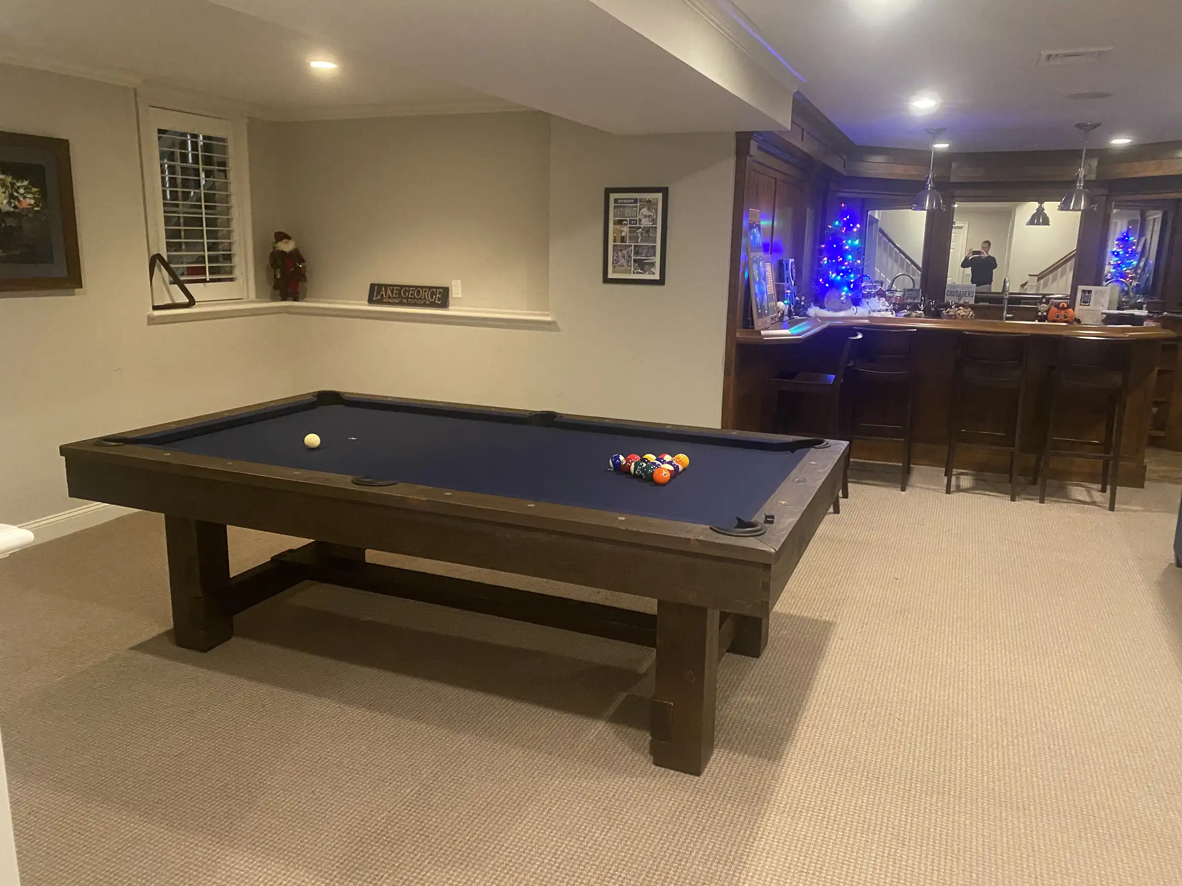 Brunswick Billiards pool table in a home game room, featuring a dark wood finish, blue felt, and ball return system. Warm lighting highlights the bar area in the background.