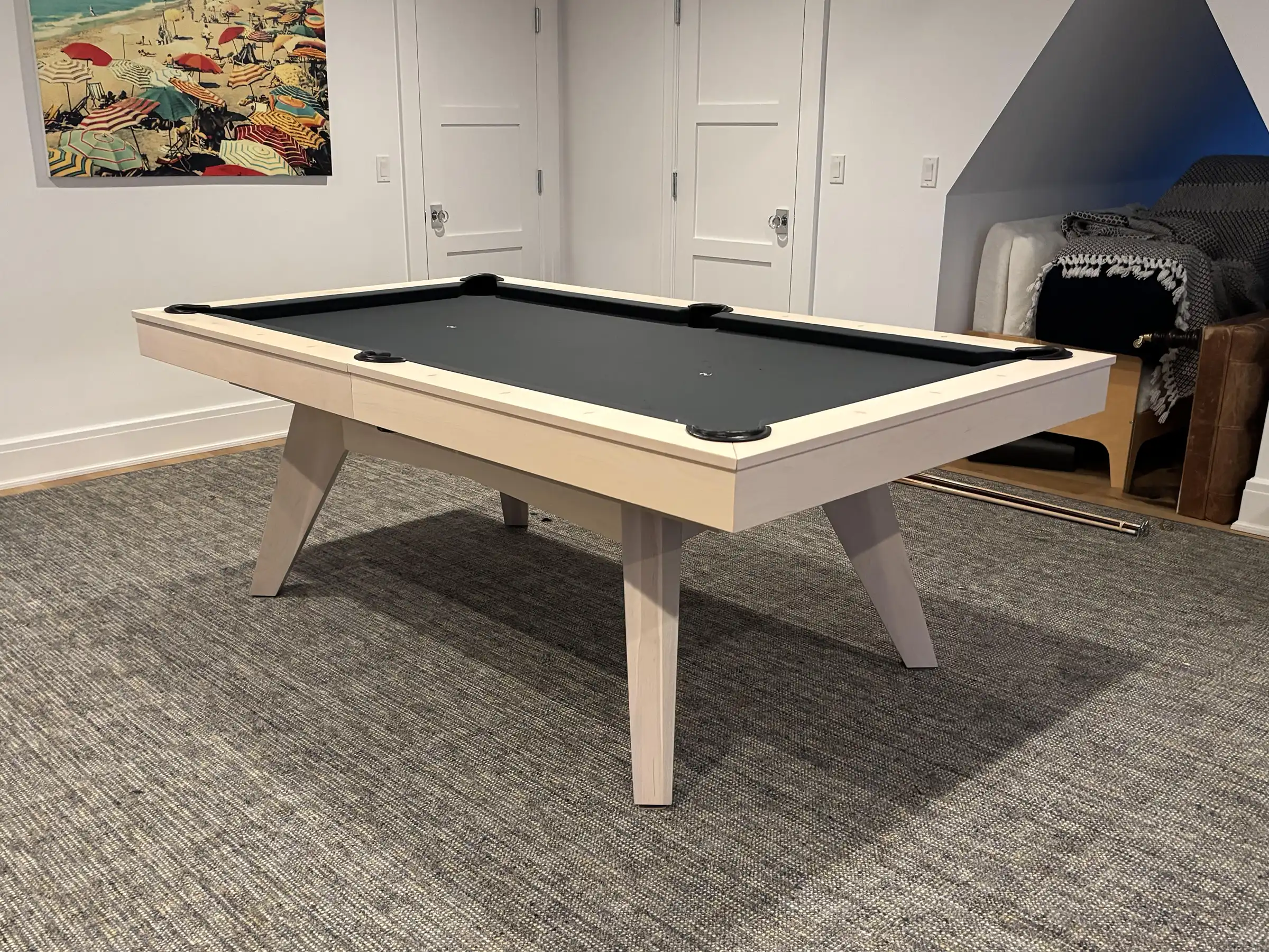 Modern pool table with light wood frame and angled legs, black felt surface, placed on gray carpet with beach umbrella artwork on wall, in minimalist room