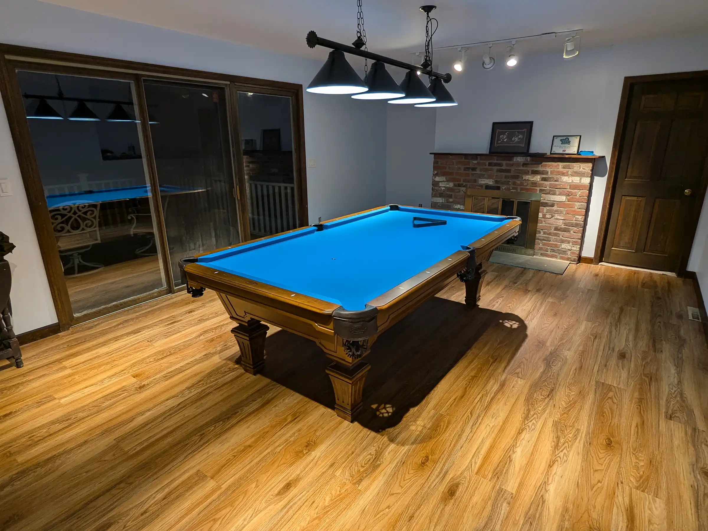Elegant home game room featuring an 8ft Olhausen pool table with vibrant blue Simonis cloth, black billiard table lighting, and brick fireplace visible. 