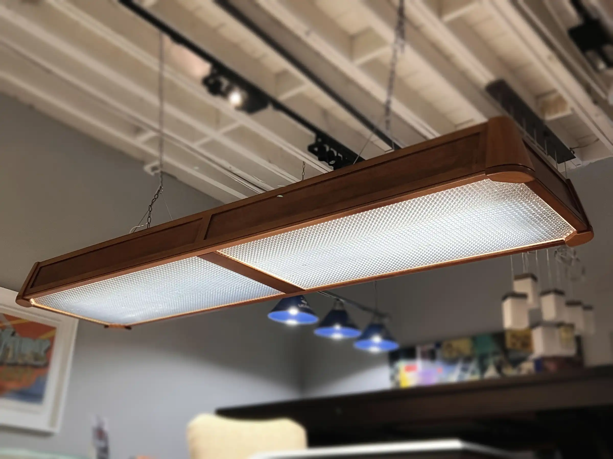 A long, rectangular billiard light fixture with a rich brown wooden frame and two frosted glass panels hangs from a white ceiling. Below, three blue pendant lights are visible, casting a soft glow. The scene is slightly blurred, suggesting a focus on the main light fixture.