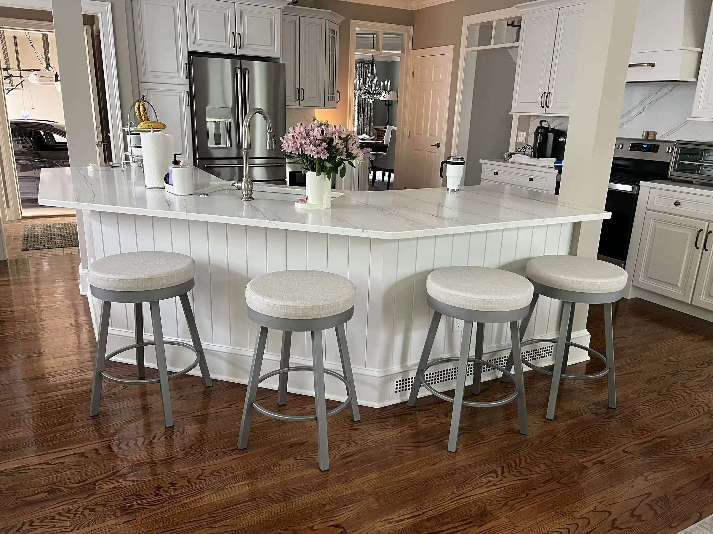 Grey counter stools with light seats line a white kitchen island in a bright kitchen with white cabinets, wood floors, and steel appliances