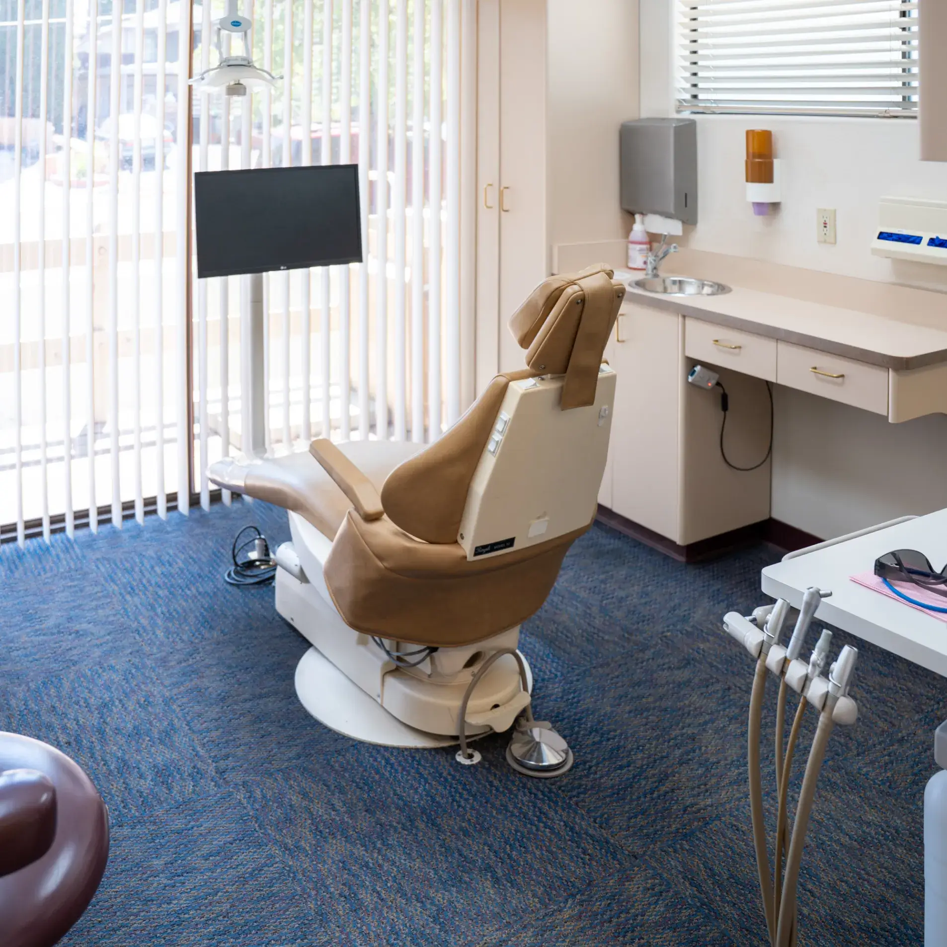 An empty dentist chair in a clean, well-lit examination room with dental tools and a monitor nearby.