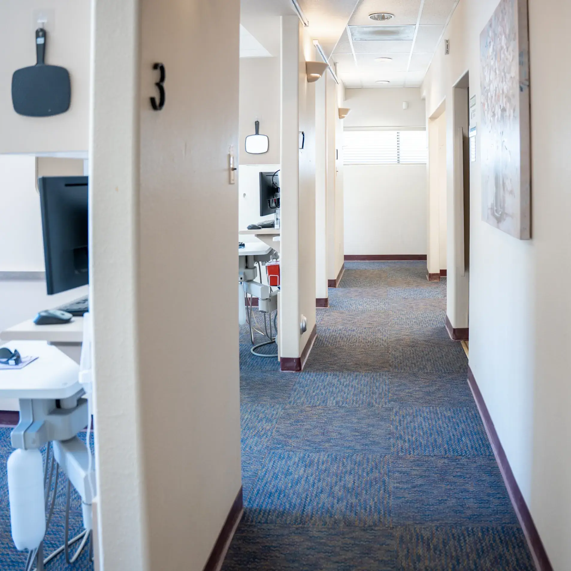 A hallway with office cubicles and computer monitors on desks, numbered doors line the corridor.