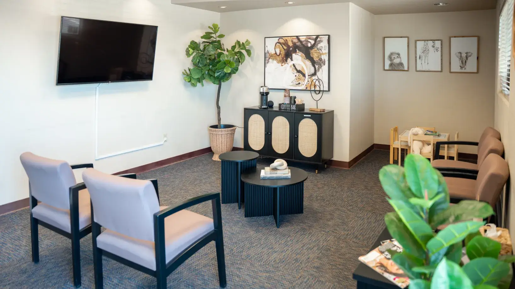 A small, modern waiting room features chairs, a TV, plants, and a coffee station.