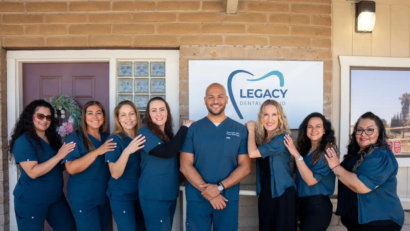 A group of eight dental clinic staff members stand smiling in front of the clinic's sign.