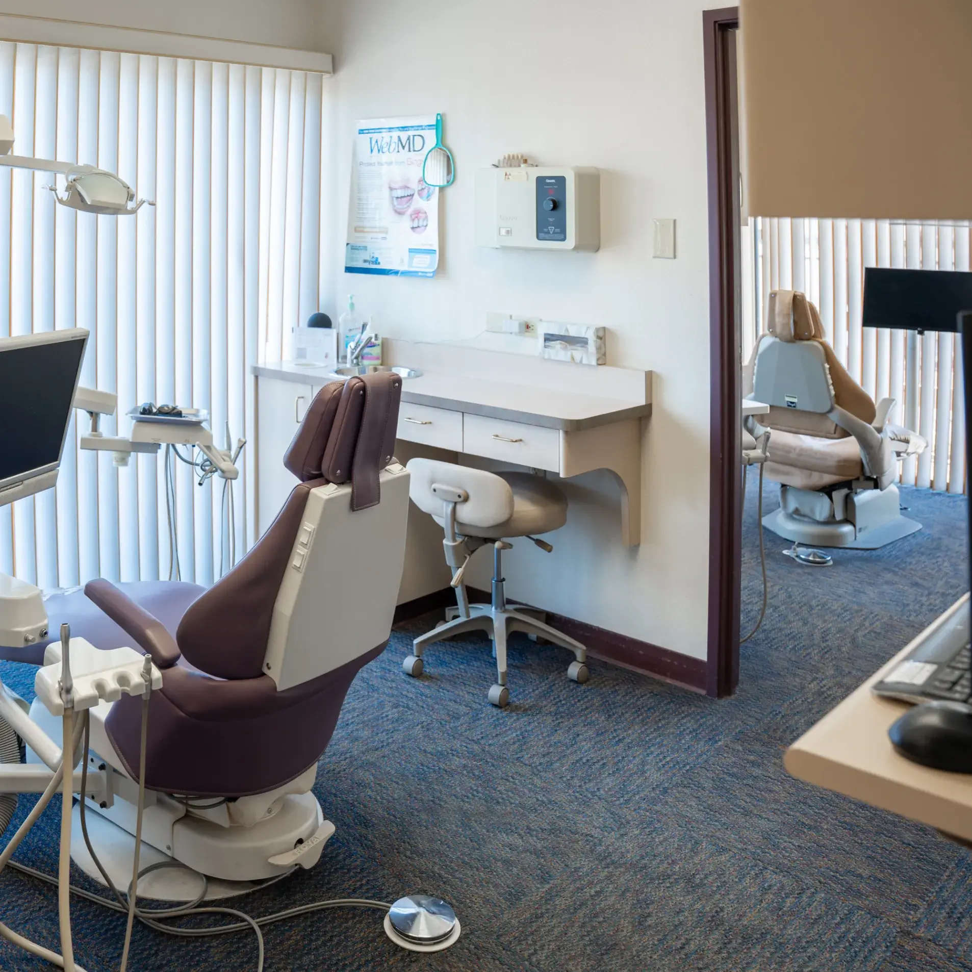 Two empty dental chairs in a clinic with equipment and blinds on the windows.