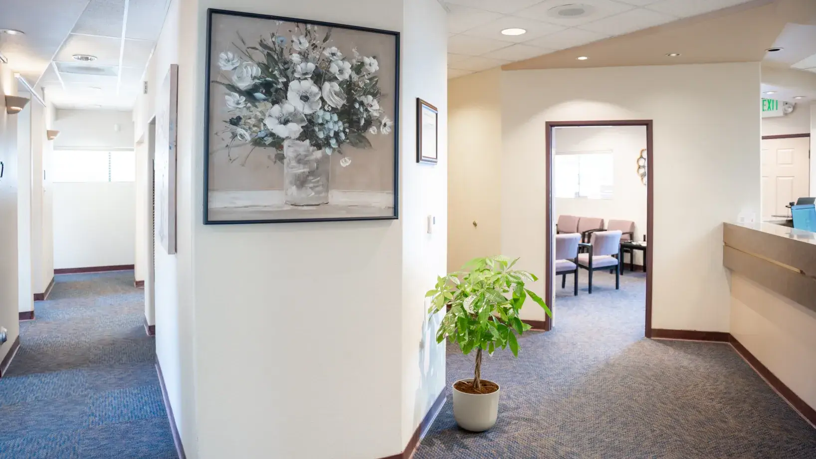 A hallway with blue carpet, featuring a potted plant and a large flower painting on the wall.