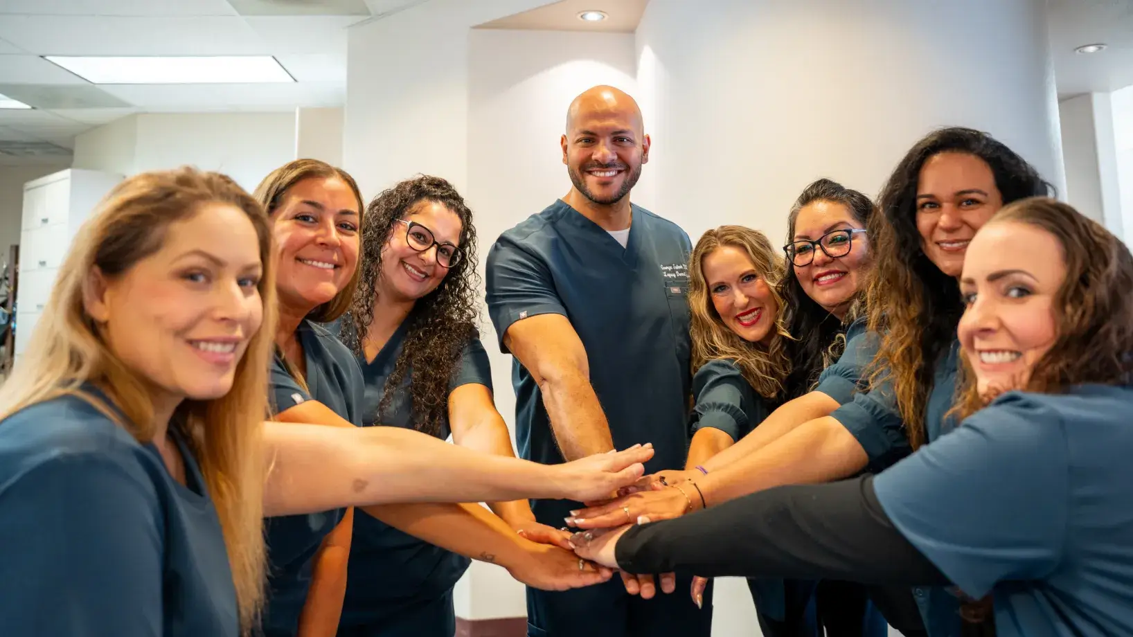 A group of smiling medical professionals in uniforms standing in a circle, stacking their hands together.