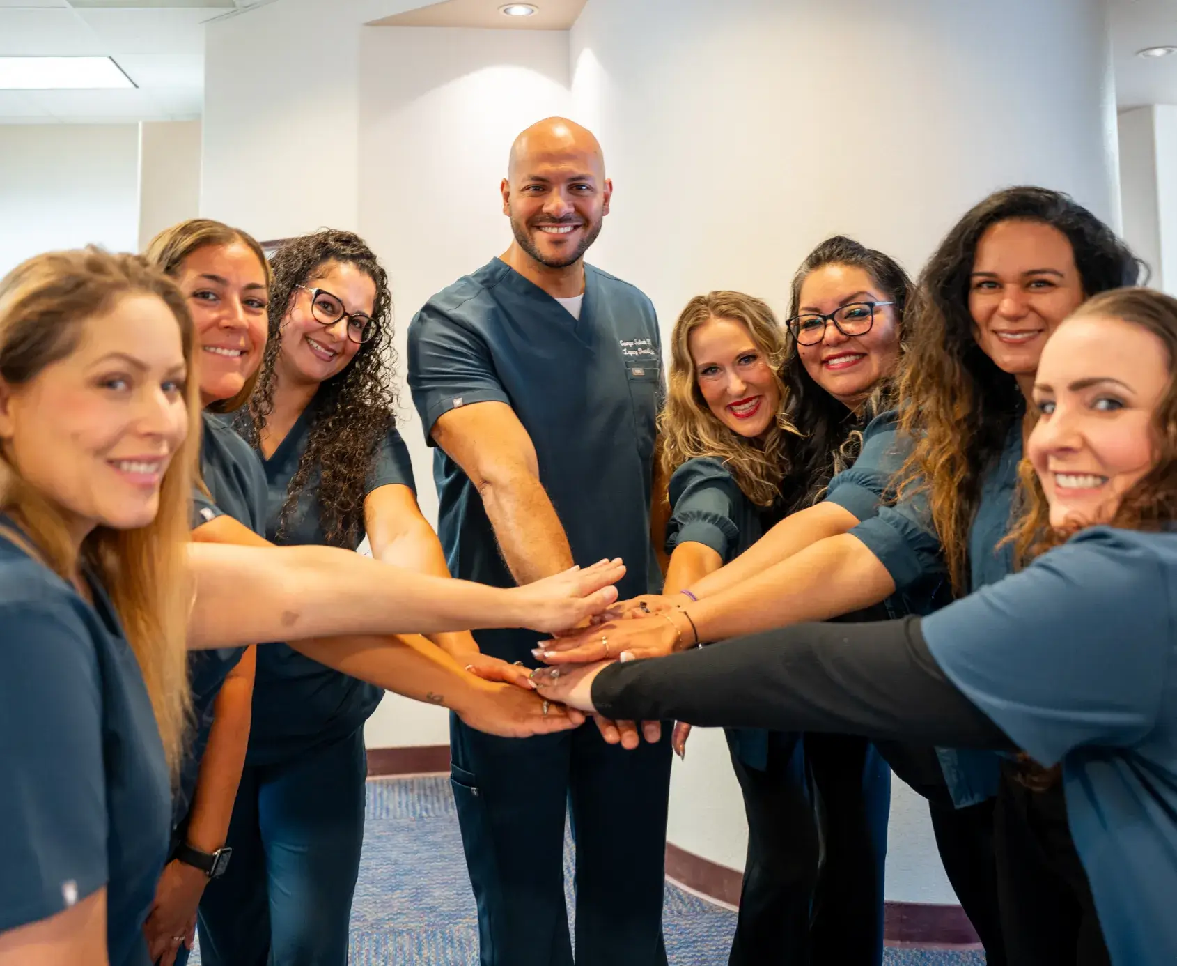 A group of people wearing medical scrubs stand in a circle, placing their hands together.