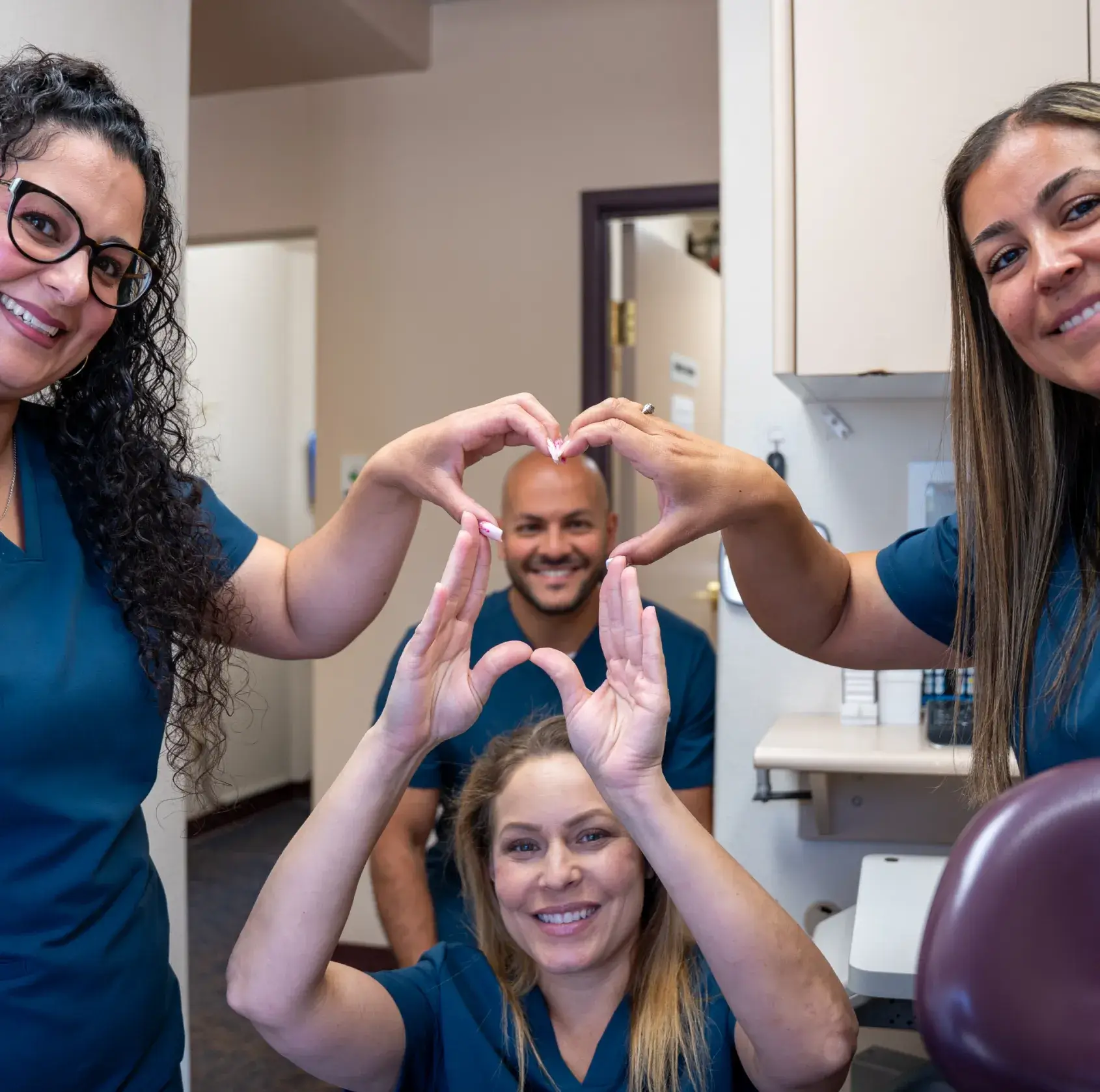 Four smiling people in scrubs form heart shapes with their hands in a dental office.