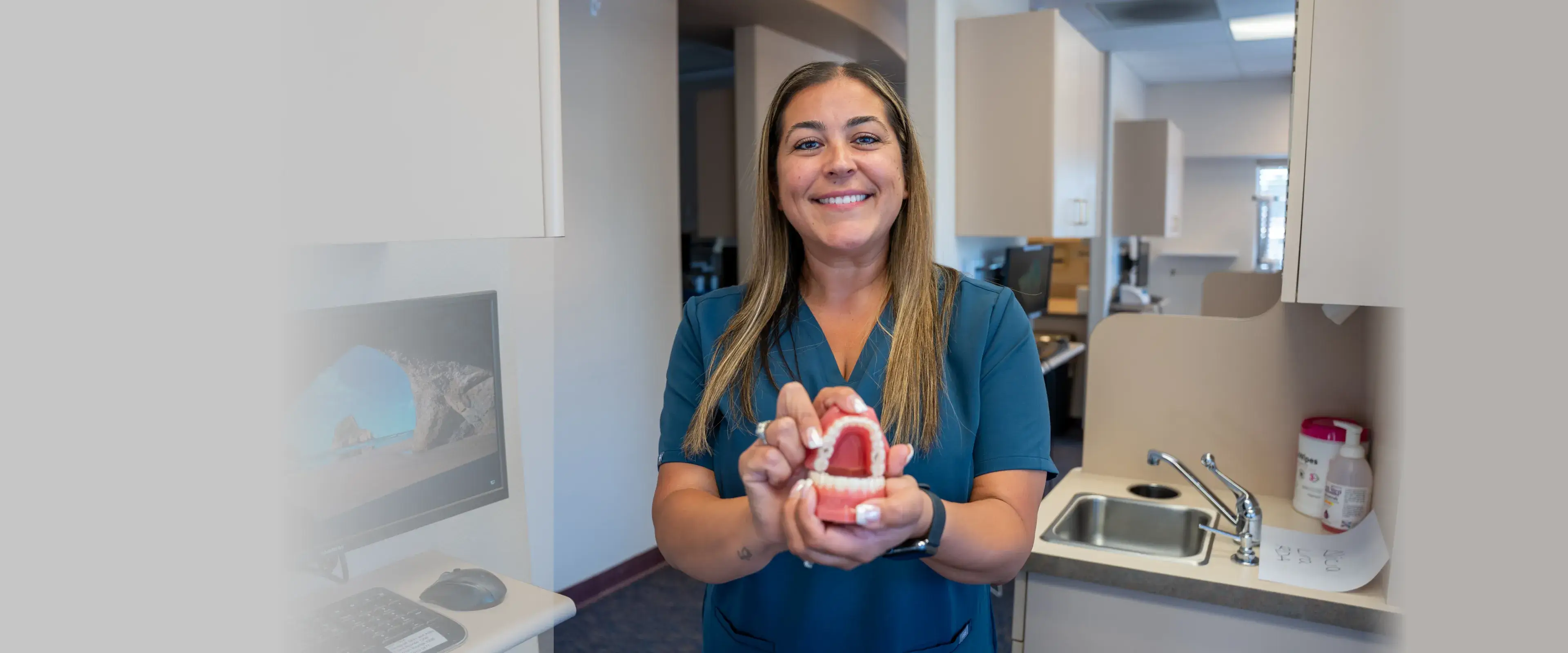 A smiling dentist holds a dental model, standing in a clinic near a computer and sink.
