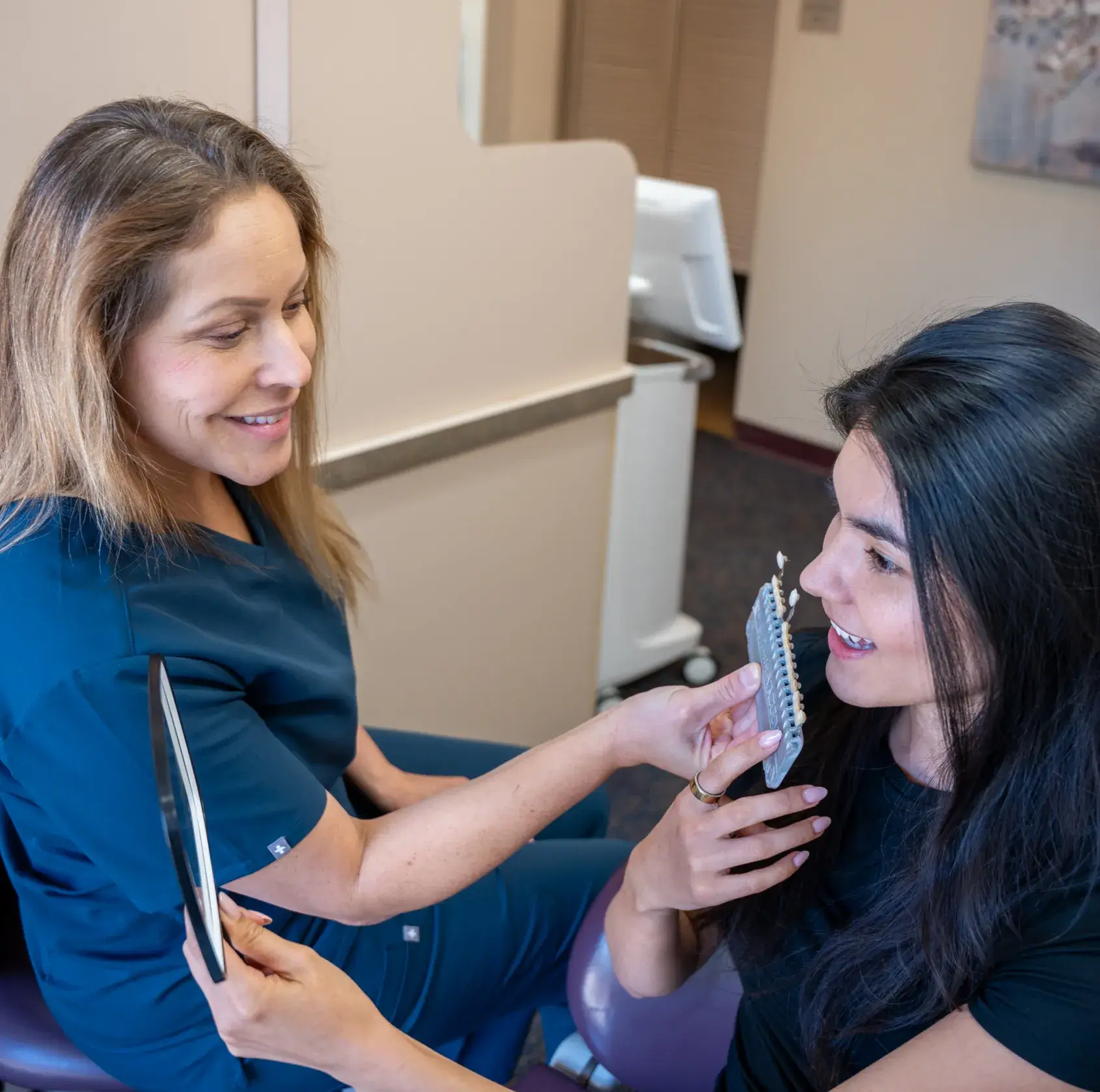 A dental professional shows a shade guide to a patient, who holds a mirror and smiles.
