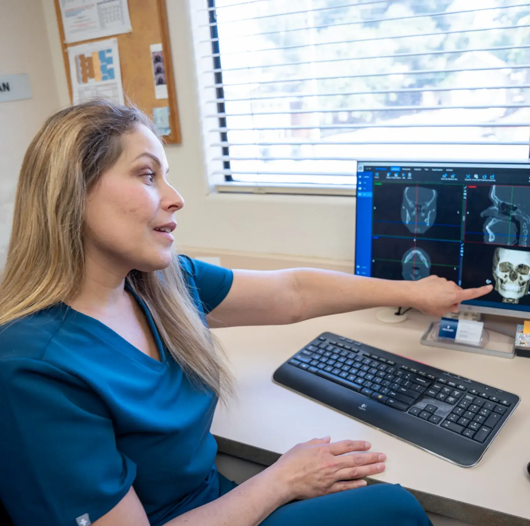 A woman in scrubs points at a skull image on a computer screen in an office.