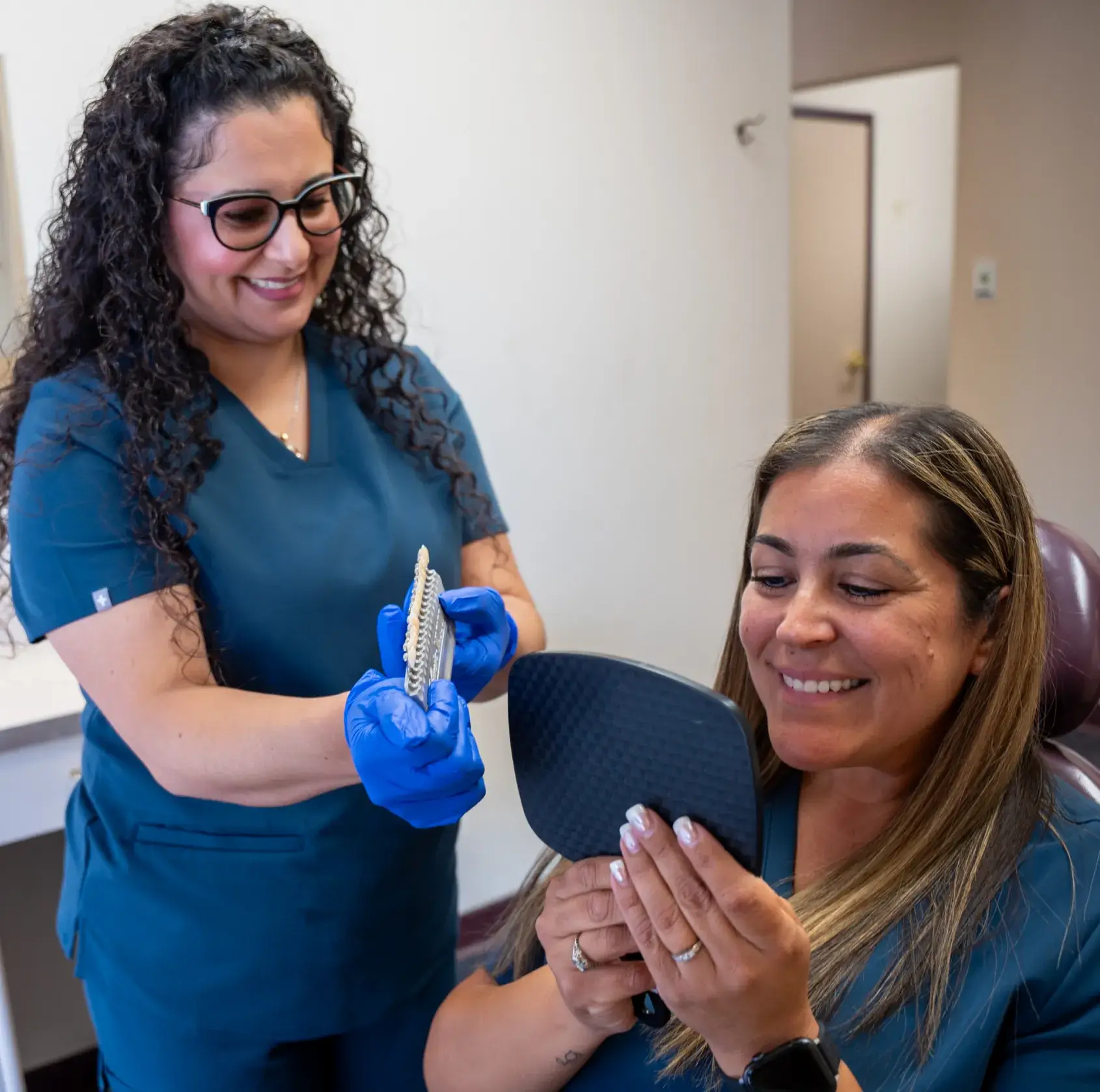 A dental professional shows a patient a dental model while the patient examines her smile in a mirror.