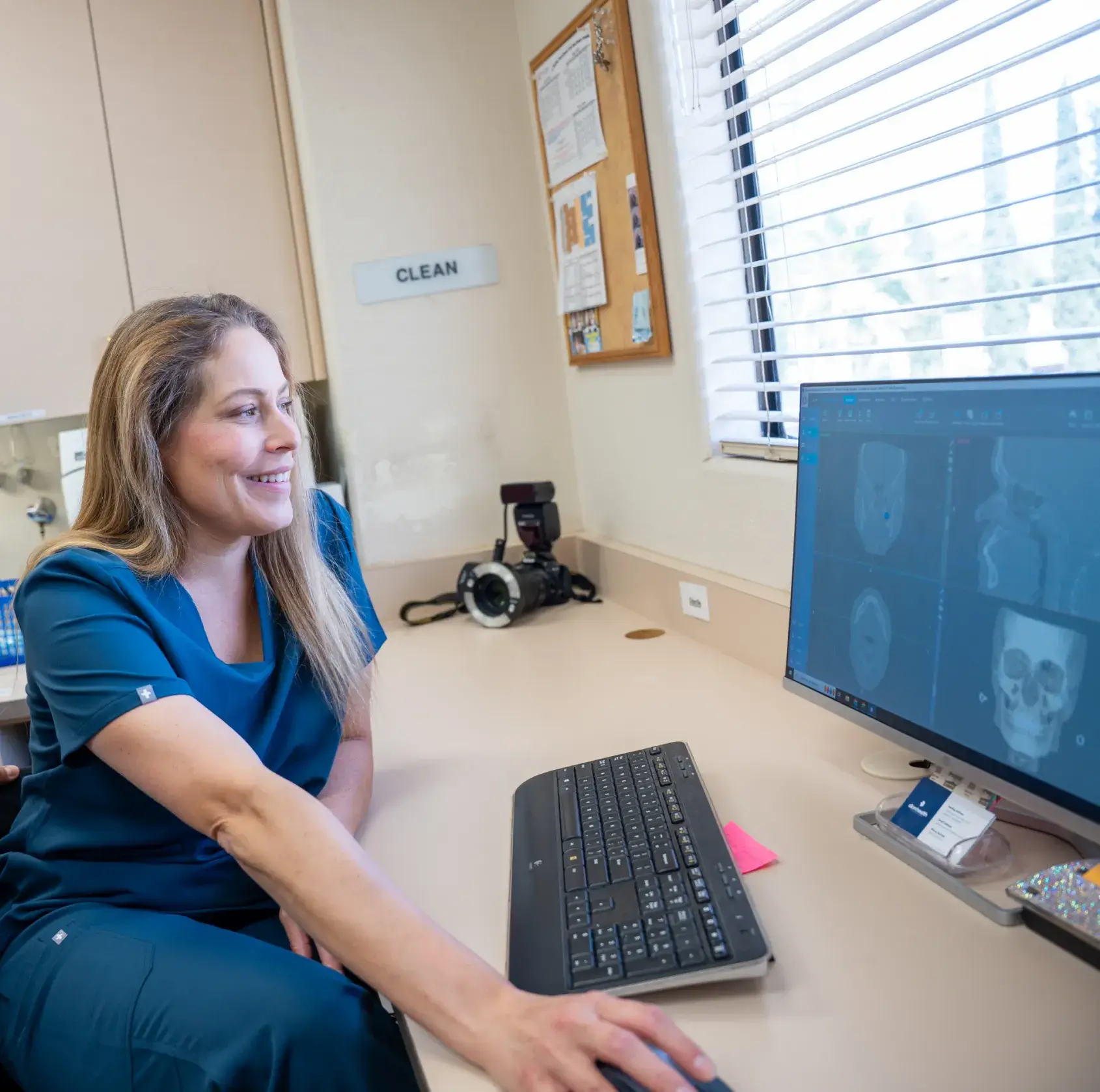 A woman in scrubs sits at a desk, looking at scans on a computer screen.