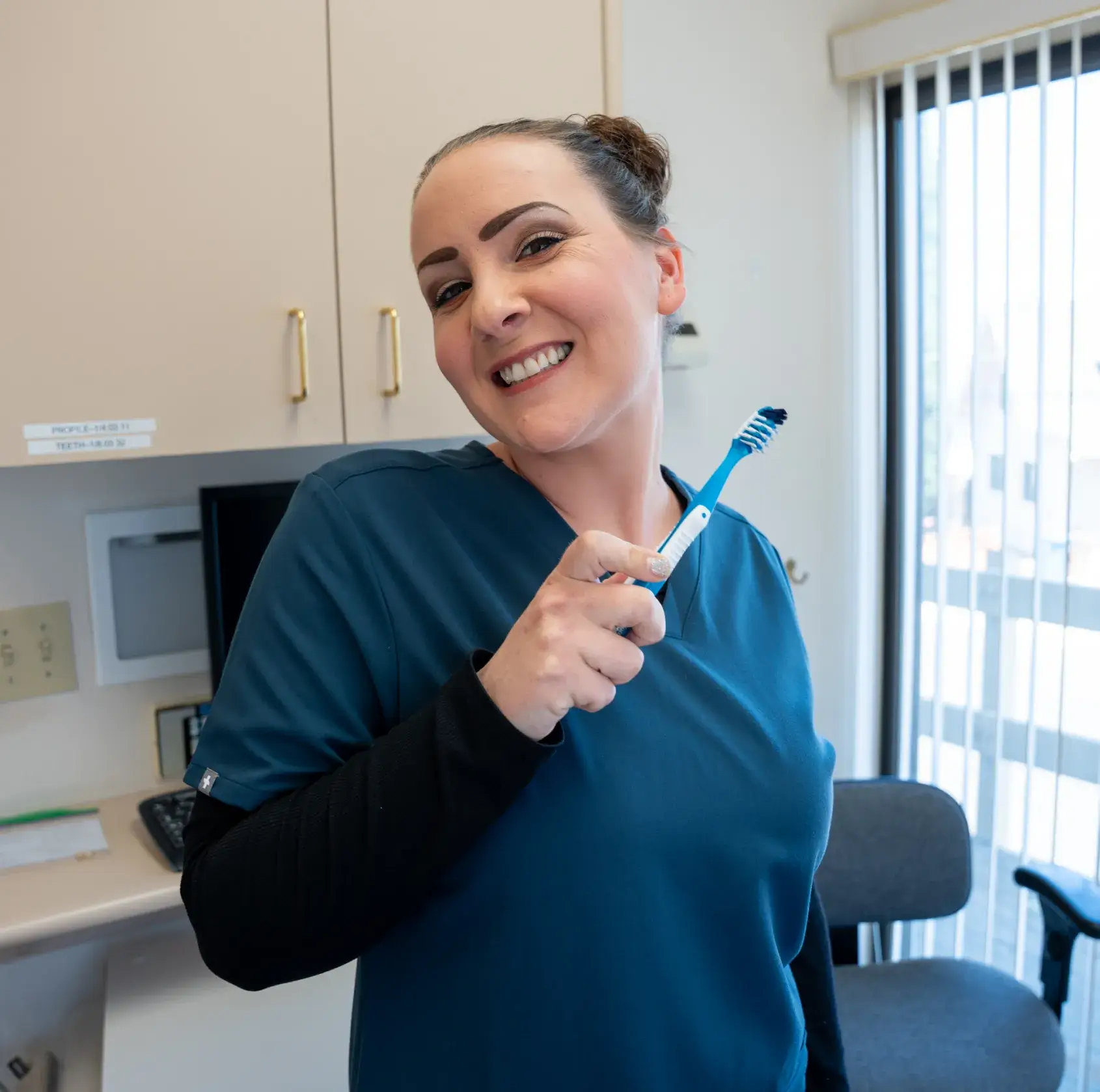 Woman in a blue scrub smiling and holding a toothbrush in a dental office.