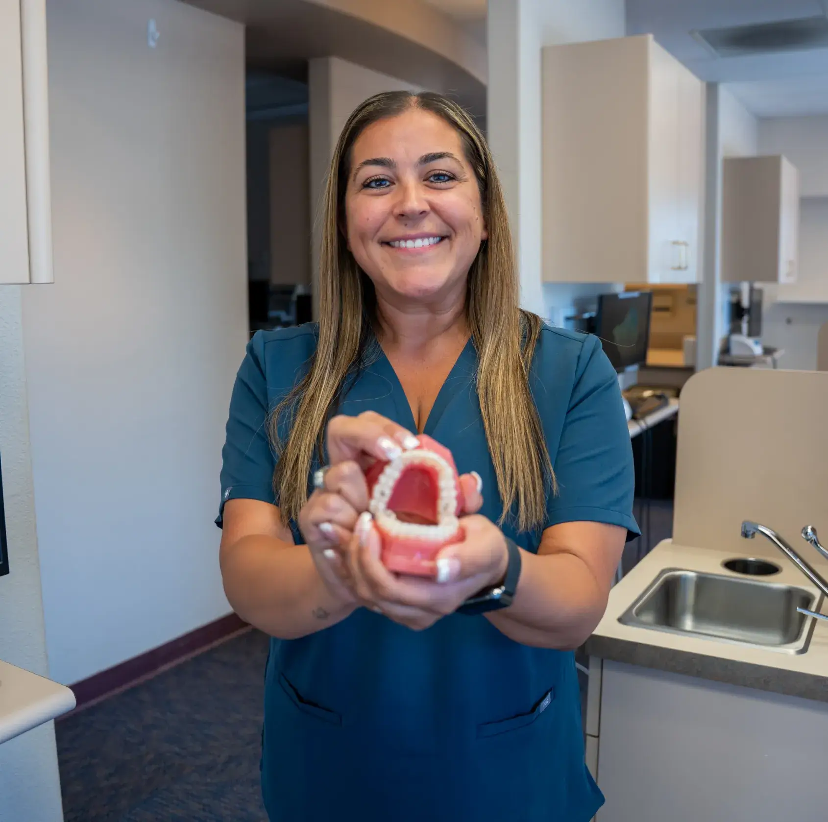 A woman in a blue uniform smiles while holding a dental model in a clinic setting.