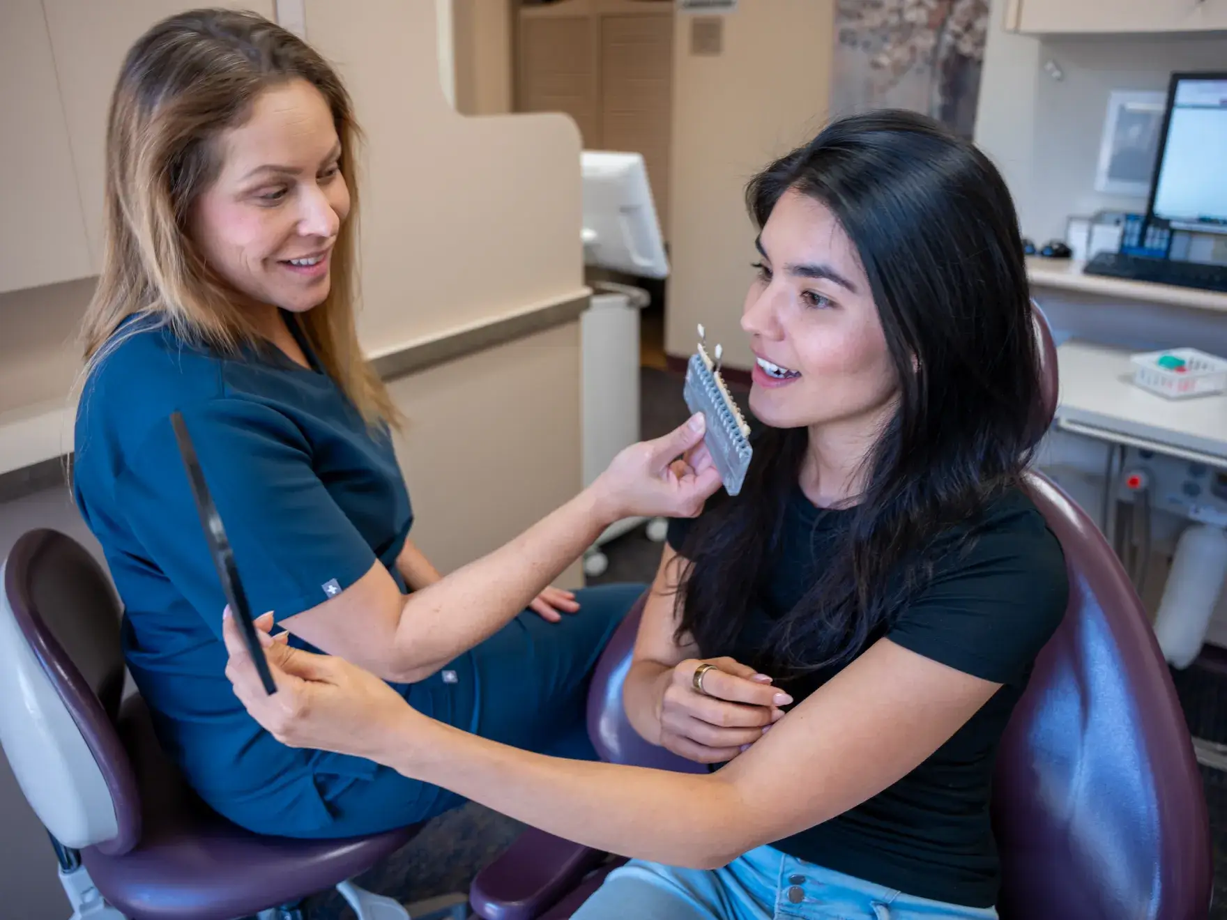 A dentist shows a patient a tooth color chart as the patient looks in a hand mirror.