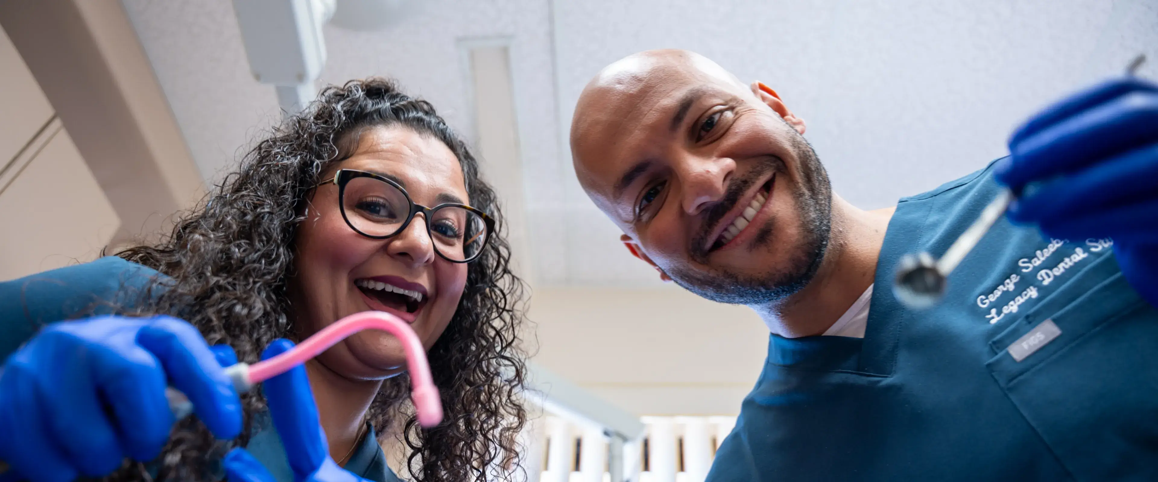 Two dental professionals smiling, wearing gloves and holding dental tools, looking down at the camera.