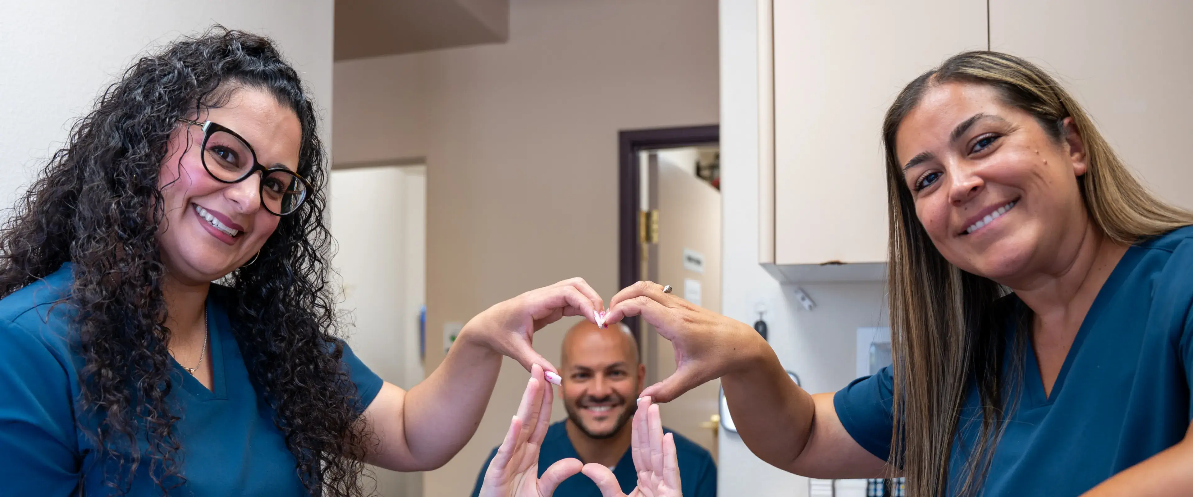 Three people in blue uniforms form a heart shape with their hands and smile at the camera.