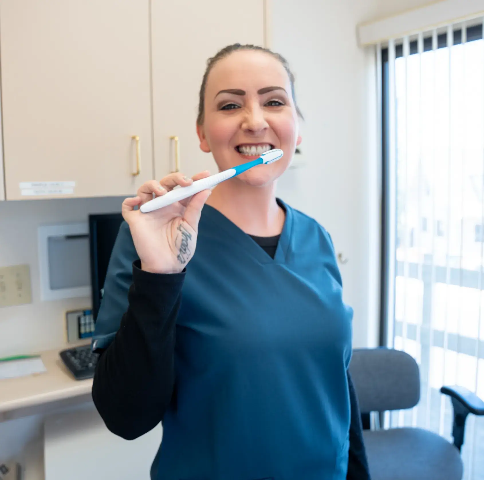 Person in blue scrubs smiling while holding a toothbrush in a dental office setting.
