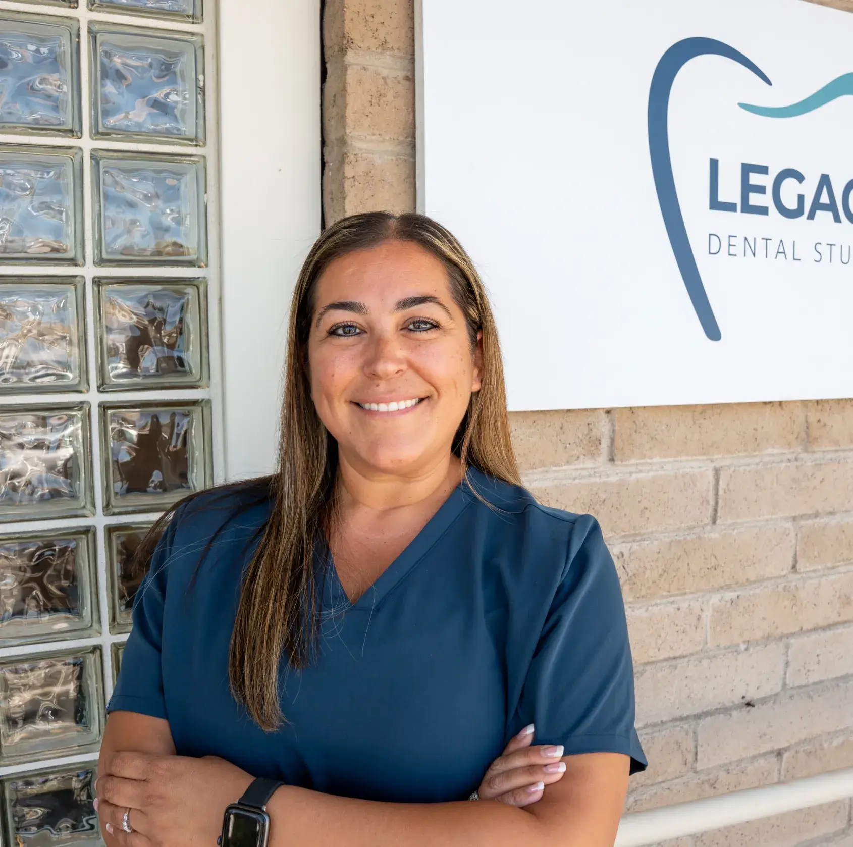 Person in blue scrubs stands smiling in front of a dental office sign.