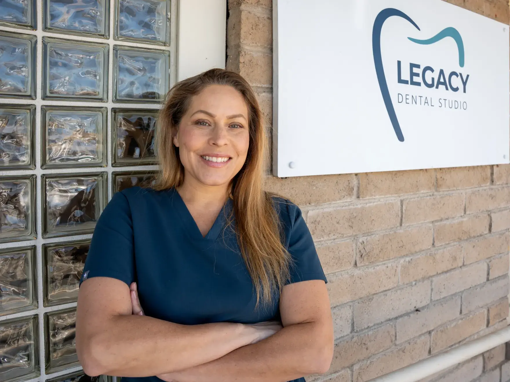 A dental professional in blue scrubs stands smiling in front of a "Legacy Dental Studio" sign.