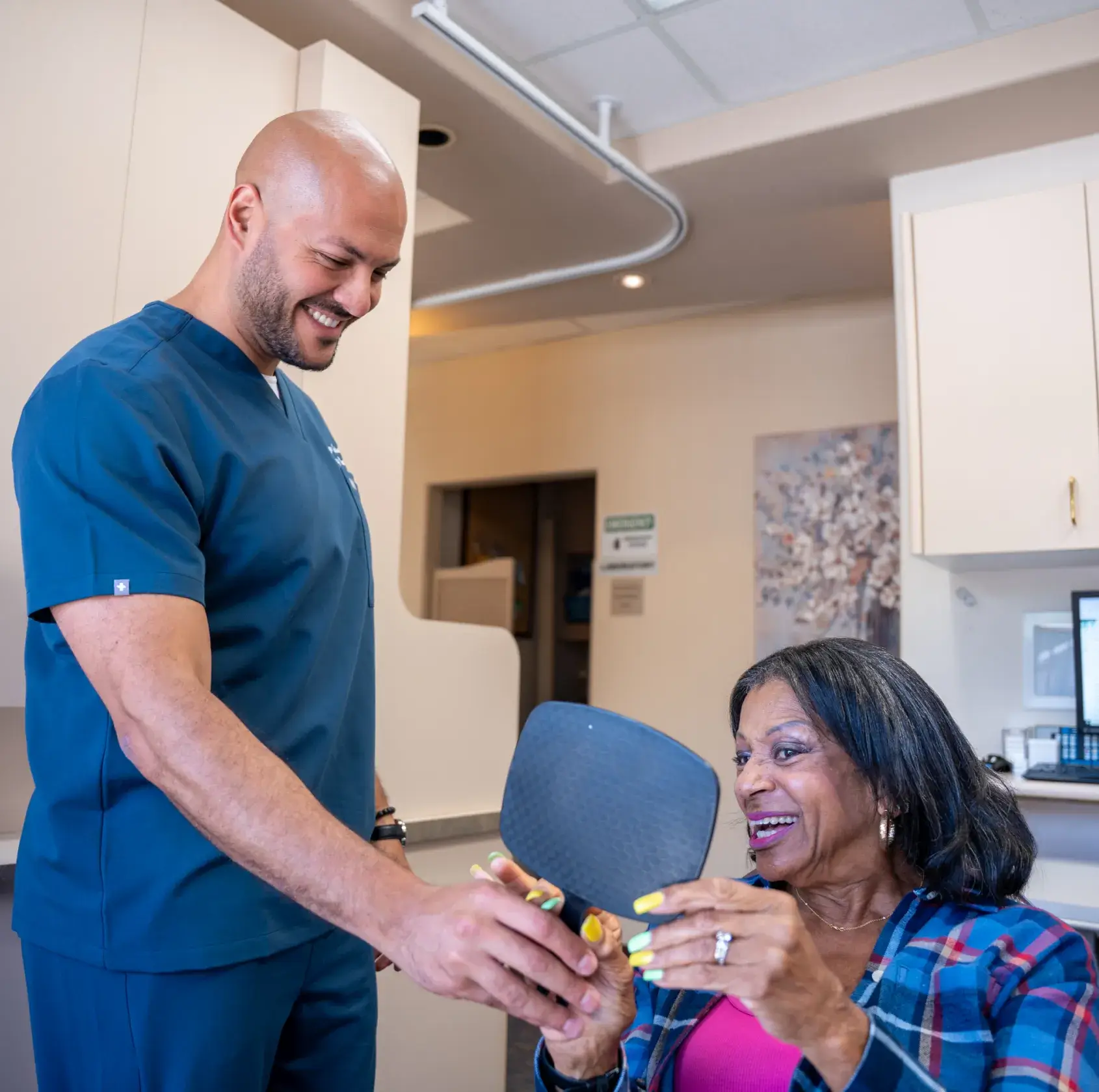 A man in scrubs shows a handheld mirror to a smiling woman in a plaid shirt.