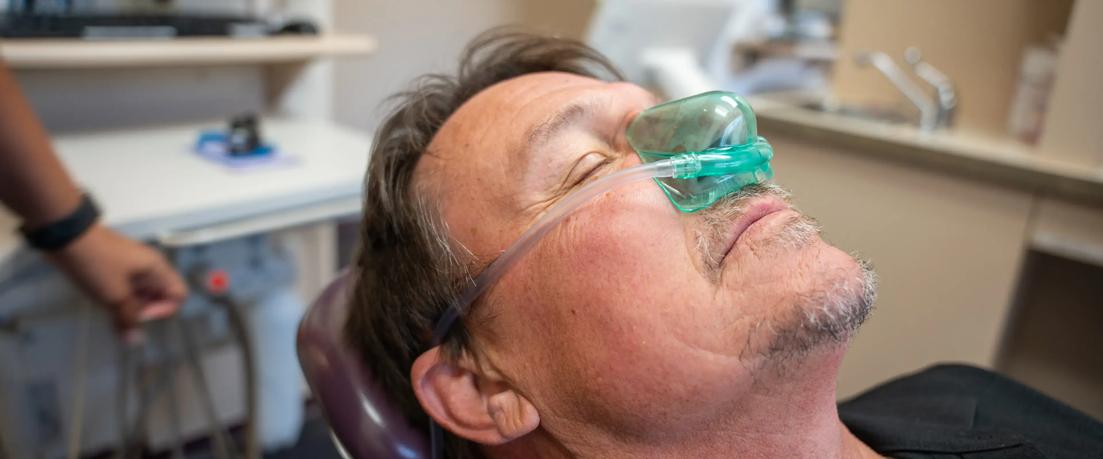 A man relaxes in a dental chair, wearing a green nitrous oxide mask over his nose.
