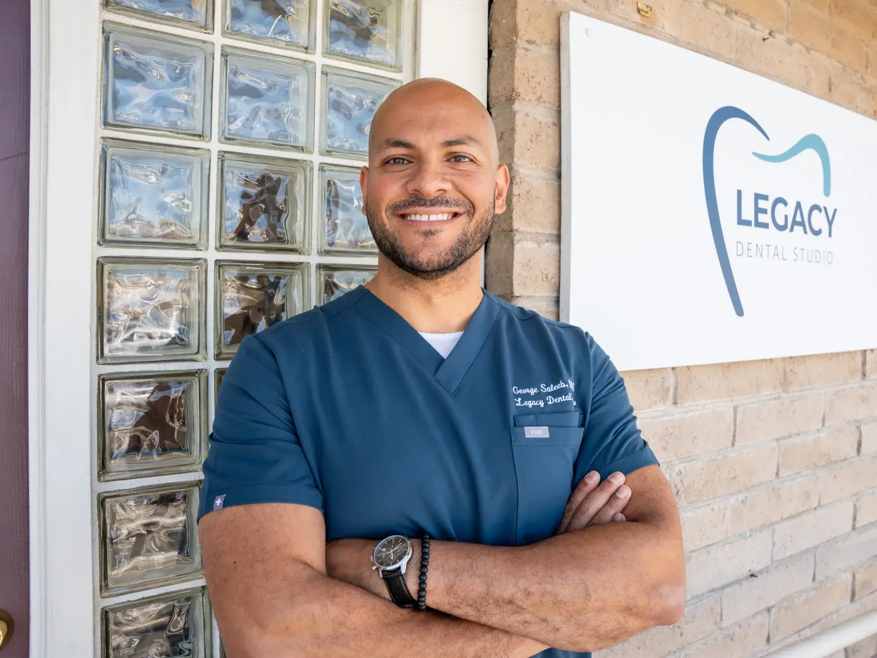 A smiling man in dental scrubs stands with arms crossed near a "Legacy Dental Studio" sign.