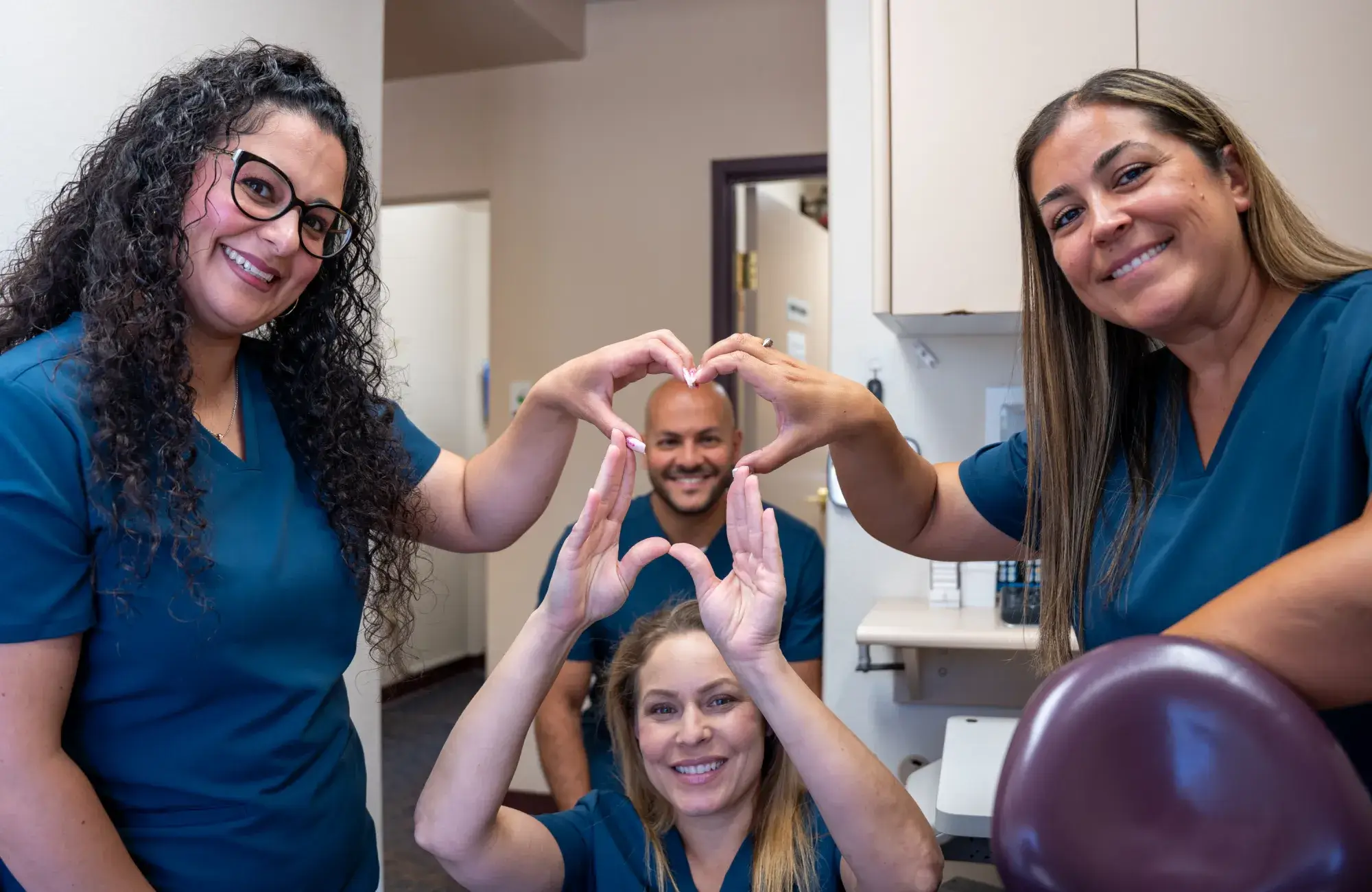 Four smiling people in blue scrubs making heart shapes with their hands in an office setting.