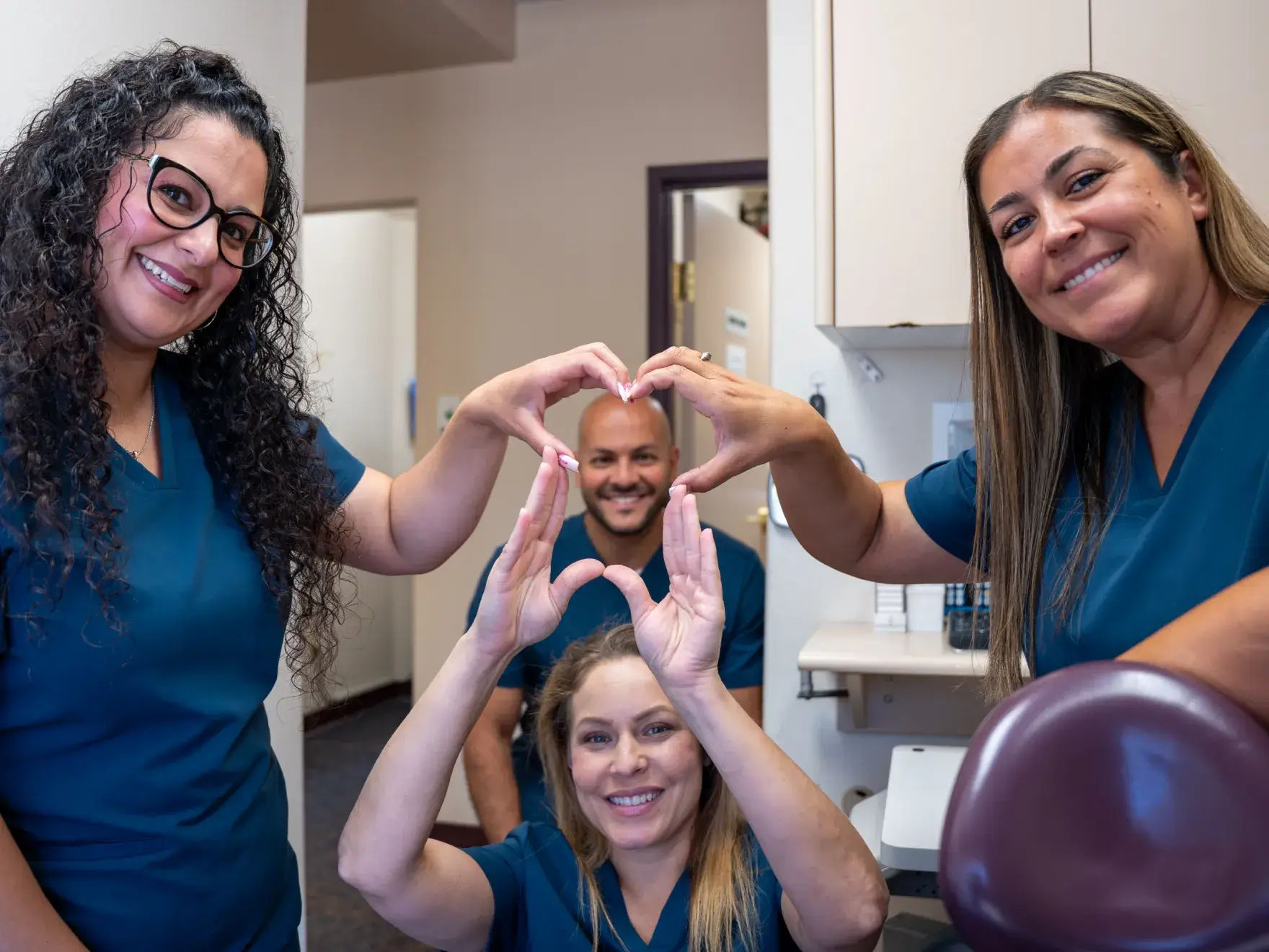 Four people in scrubs forming a heart shape with their hands, smiling in a room.