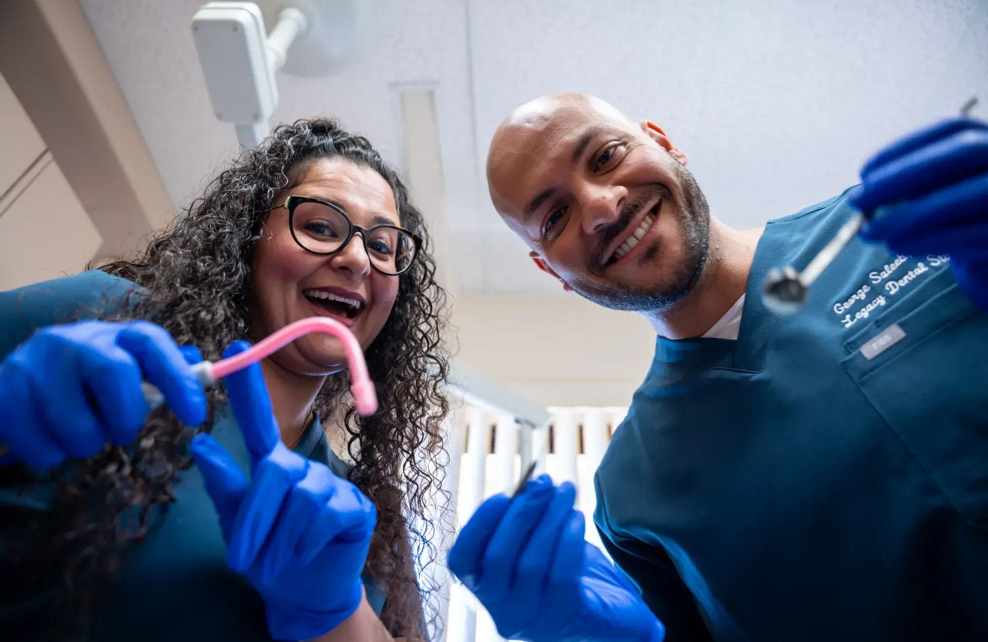 Two dentists wearing gloves smile while holding dental instruments, looking directly at the camera.