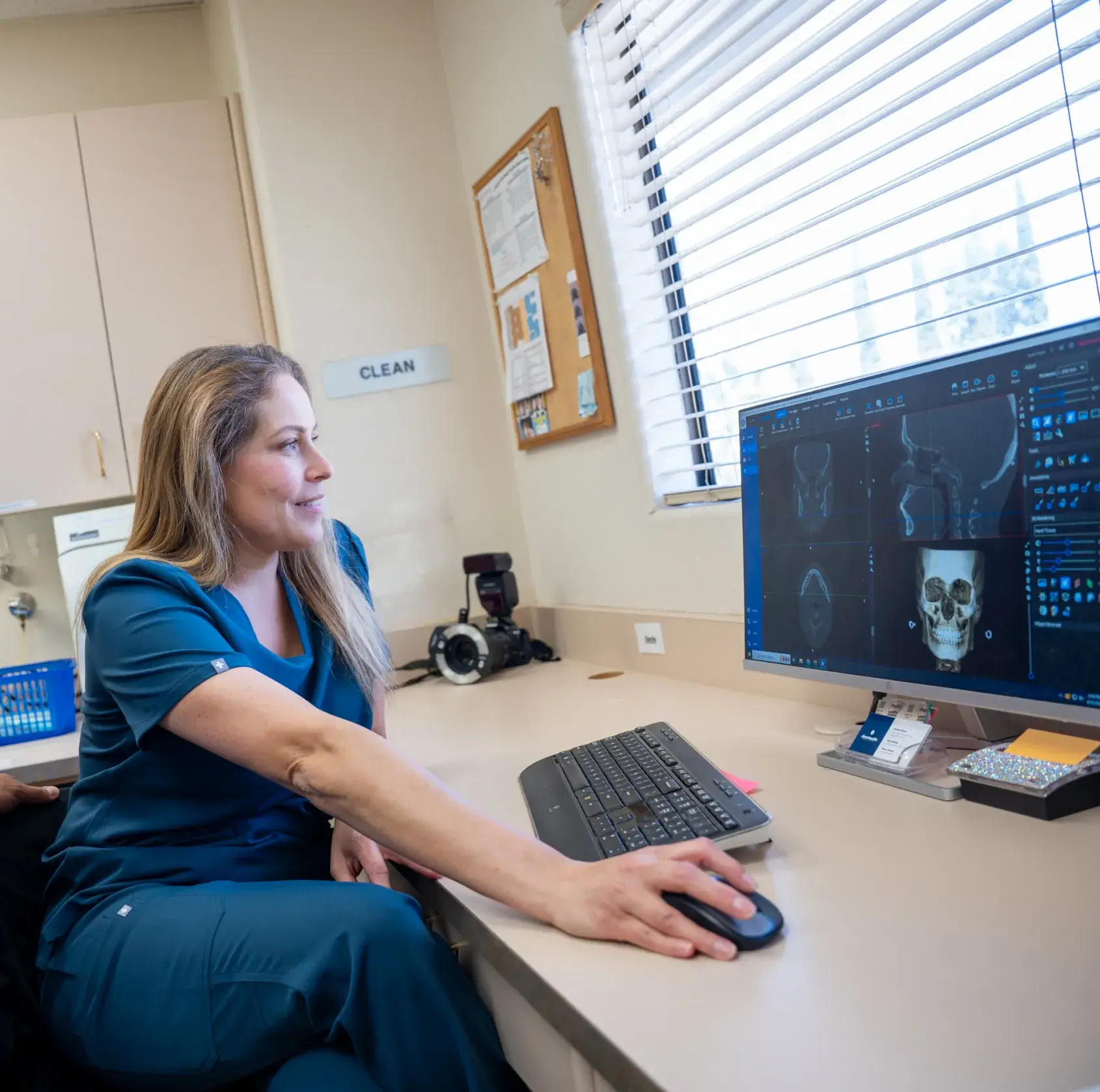 A woman in scrubs looks at skull X-rays on a computer screen in a medical office.