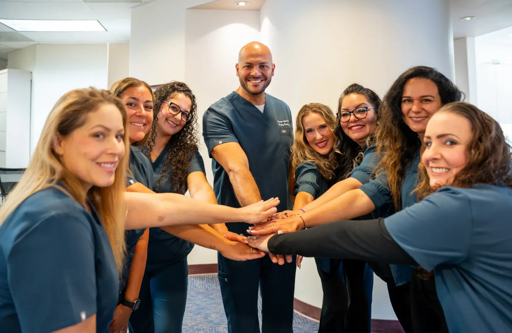 A group of people in matching scrubs smiling and placing their hands together in a circle.