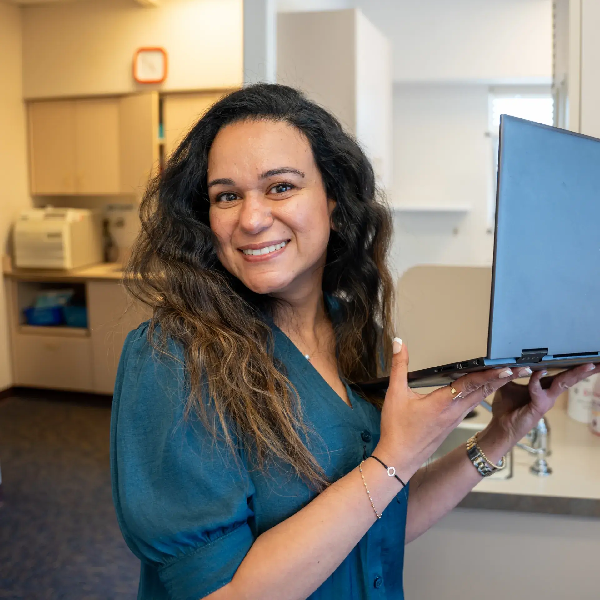 A woman with long hair smiles while holding an open laptop in an office setting.