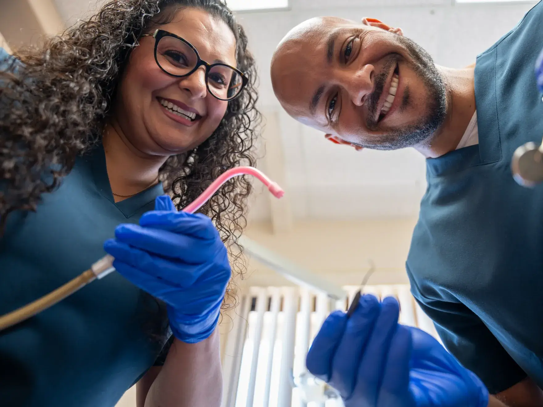 Two dentists wearing gloves and scrubs smile while holding dental tools, looking down at a patient.