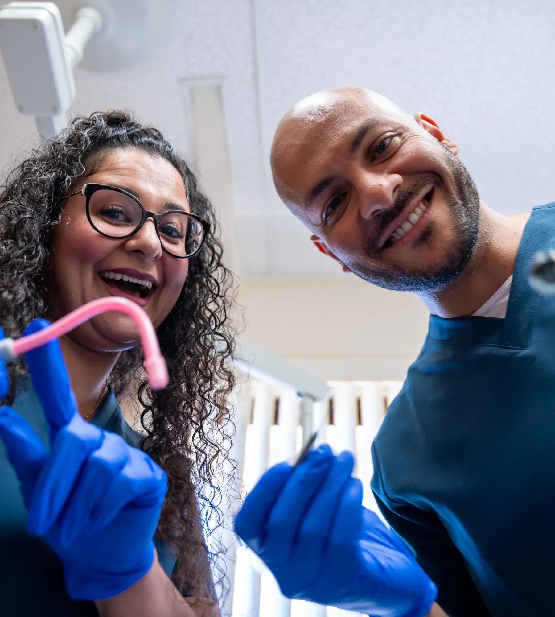 Two dentists in blue gloves smiling and holding dental tools, looking down towards the camera.