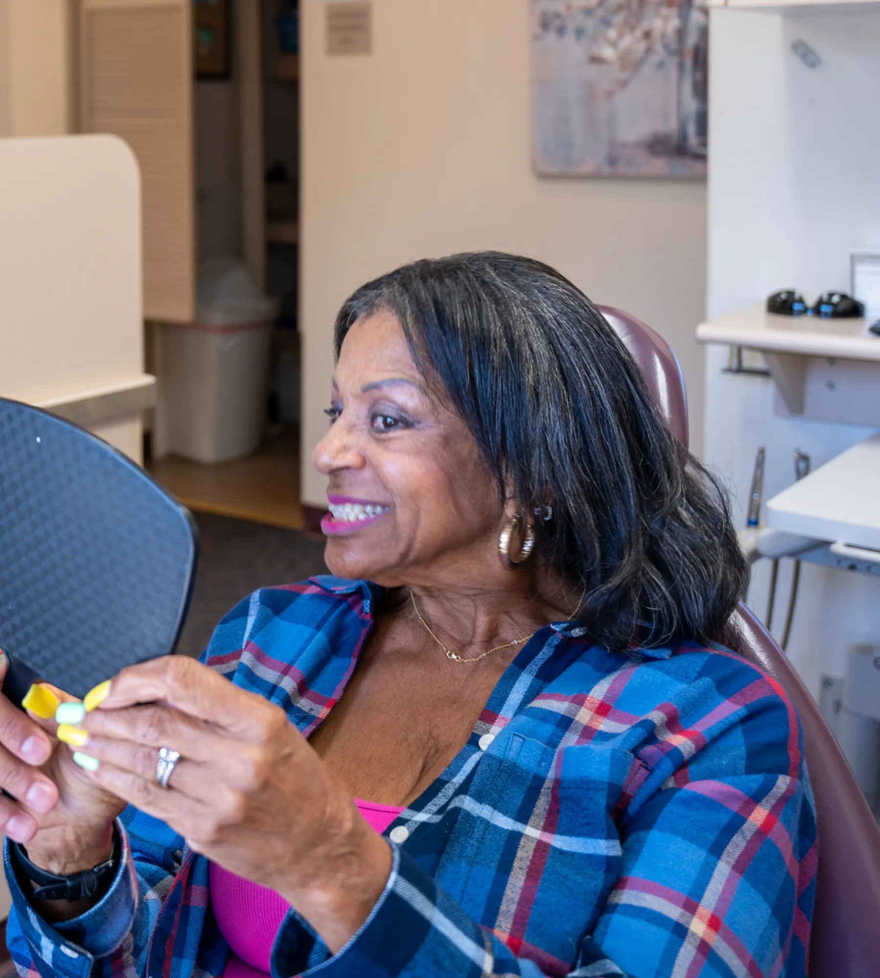 A woman in a plaid shirt smiles while looking at herself in a handheld mirror.