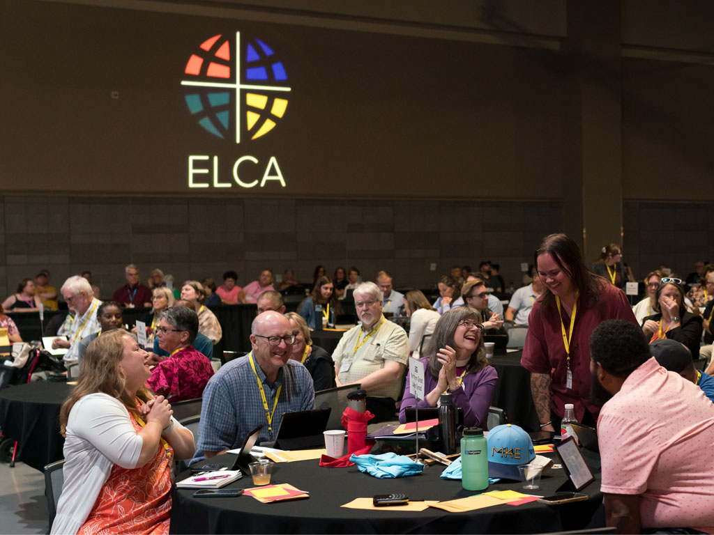  a group of people laughing around a round table in a large crowded room with the ELCA logo, consisting of a cross and four colored panels, projected on the wall behind them. 