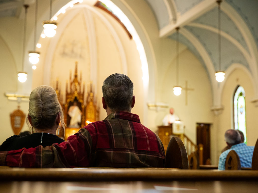 photo from a church pew looking toward the altar with a view of a man and woman with gray hair, the man wearing a red flannel with his arm around the woman as they look forward.