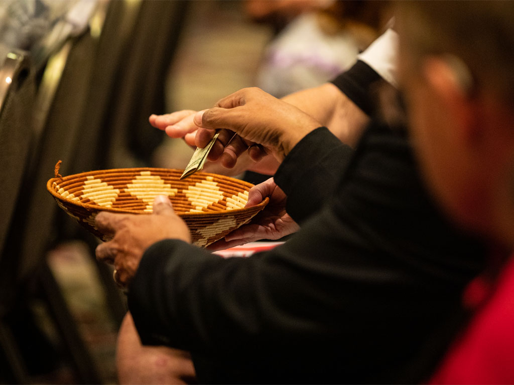 A figure in a black shirt passes a woven offering basket to the person next to them as a hand places a folded $5 bill inside  