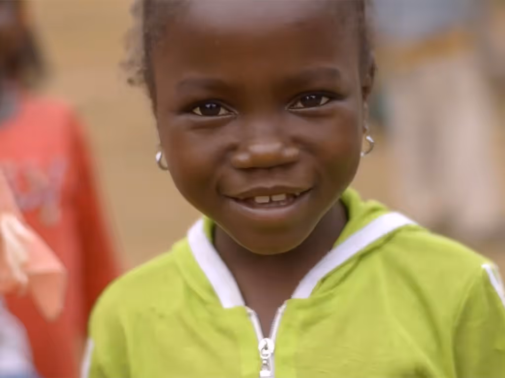 small child with dark skin, and earrings in both ears wearing a green zip-up shirt smiles for the camera