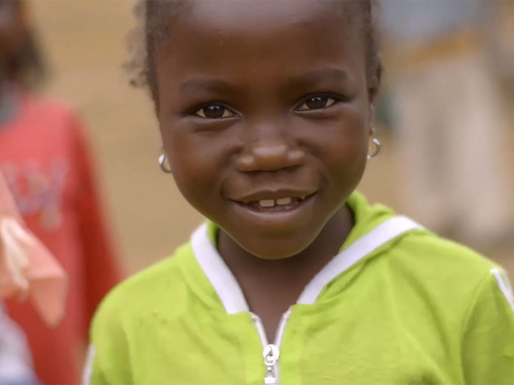 A small child with dark skin and earrings in both ears, wearing a green zip-up shirt, smiles for the camera.