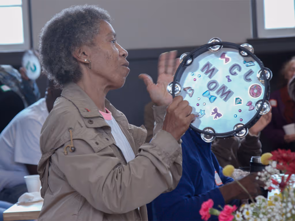 African American woman with short gray hair wearing a gray jacket plays a tambourine with the word “MOM” and various stickers of bows and bowties.