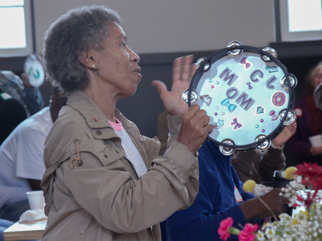 African American woman with short gray hair wearing a gray jacket plays a tambourine with the word “MOM” and various stickers of bows and bowties.