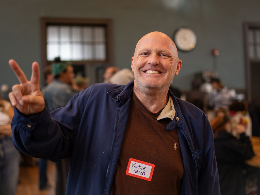 Bald man wearing a blue jacket over a brown sweater with a Polo logo giving the peace sign to the camera as he stands in front of an out-of-focus room full of people.